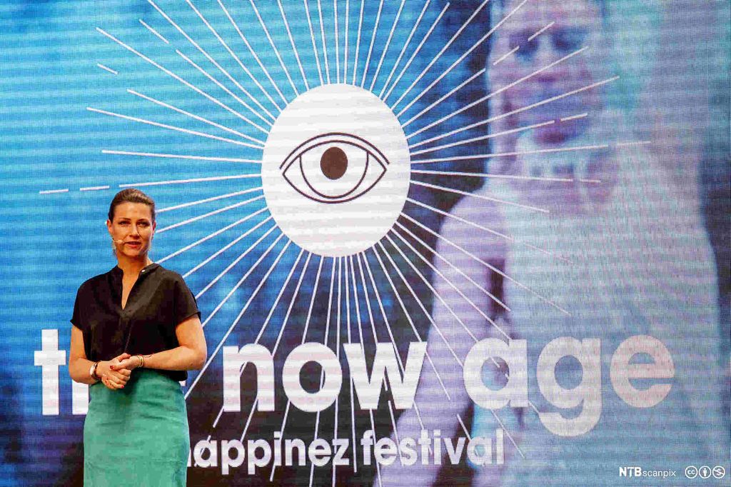 Prinsesse Märtha Louise med mikrofon. Står foran en stor skjerm med bilde av et øye med linjer som stråler ut. Under øyet står det skrevet "The Now Age. The Happinez Festival". Foto.