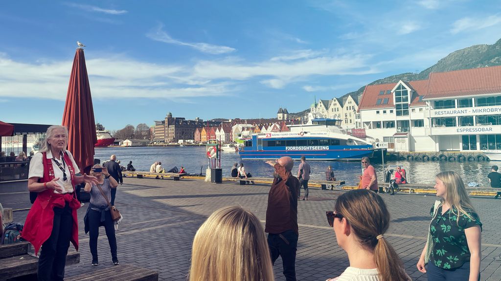 Guide står og snakkar til eit turfølge på ei brygge i ein by. Foto.
