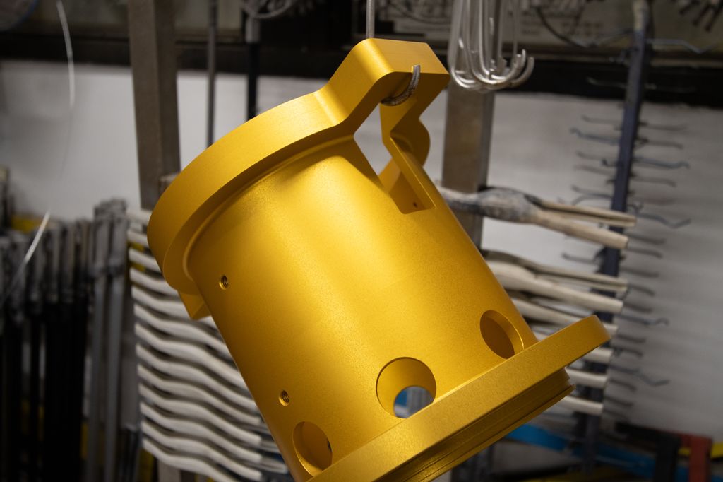 Rørstuss i aluminium som er eloksert med et gullfarga belegg. og som henger på en krok. I bakgrunnen klemmer og andre typer verktøy, satt opp i ryddige rader. Foto.