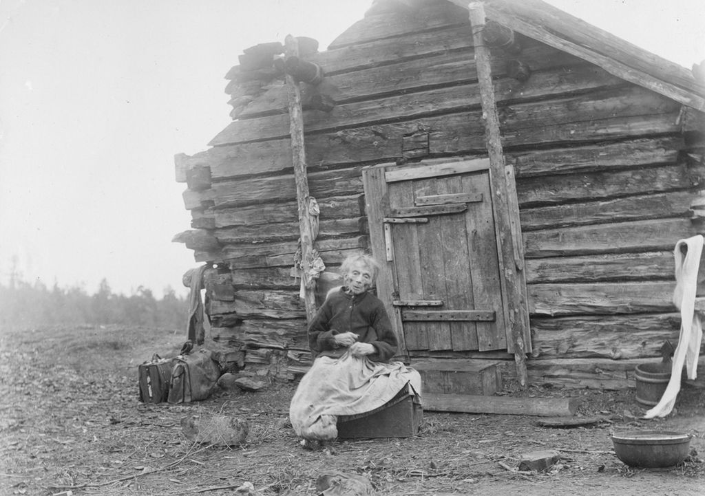 En gammel, liten kone sitter foran en koie. Hun har mørk skjorte og langt skjørt. Svart-hvitt foto.
