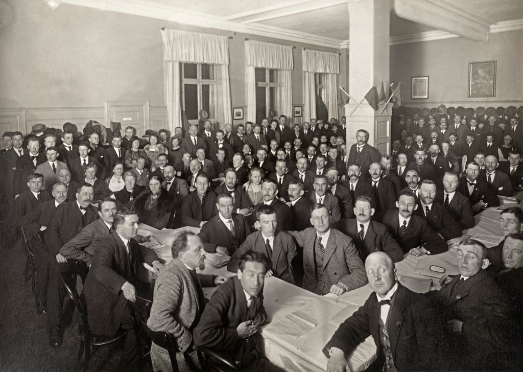 Veldig mange menn og noen få kvinner sitter rundt et langbord med hvite duker. Det står også mange langs veggene i bakgrunnen. Foto. 