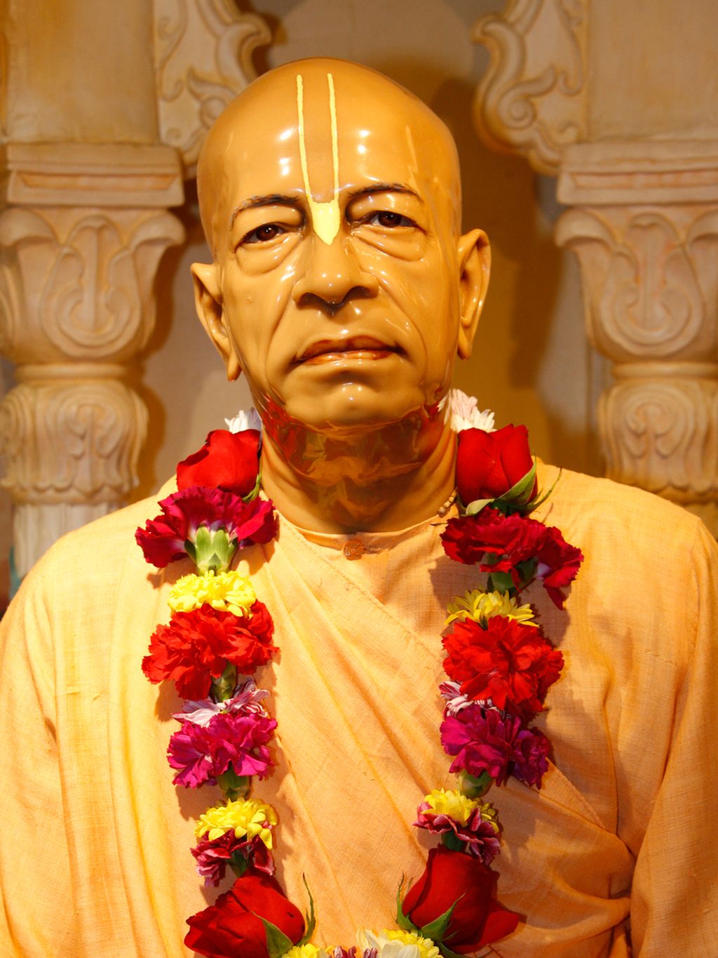 Statue av mann med oransje kjortel, bomsterkrans rundt halsen og merke i panna. Foto.