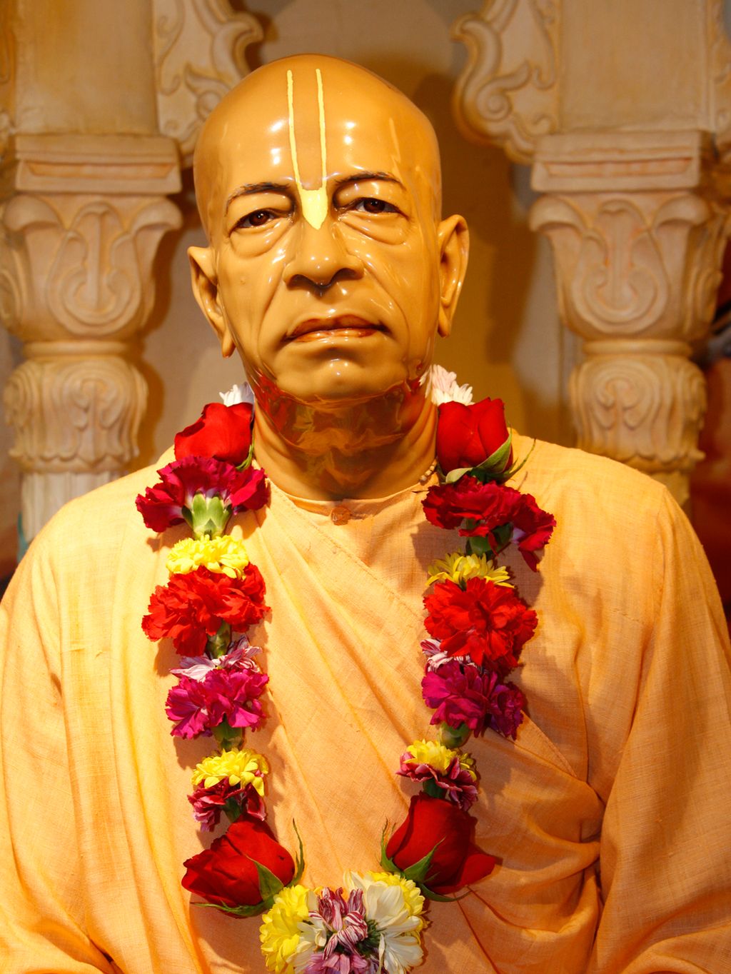 Statue av mann med oransje kjortel, bomsterkrans rundt halsen og merke i panna. Foto.