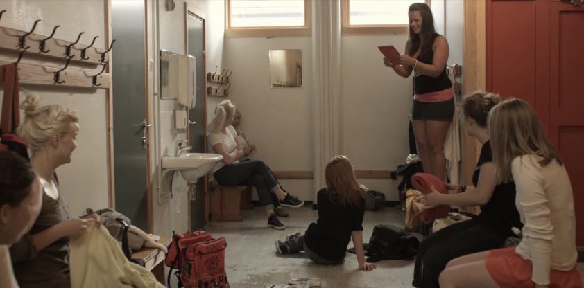 Ei jente står på en benk i jentegarderoben og leser fra ei bok. De andre jentene ser på henne. Foto.