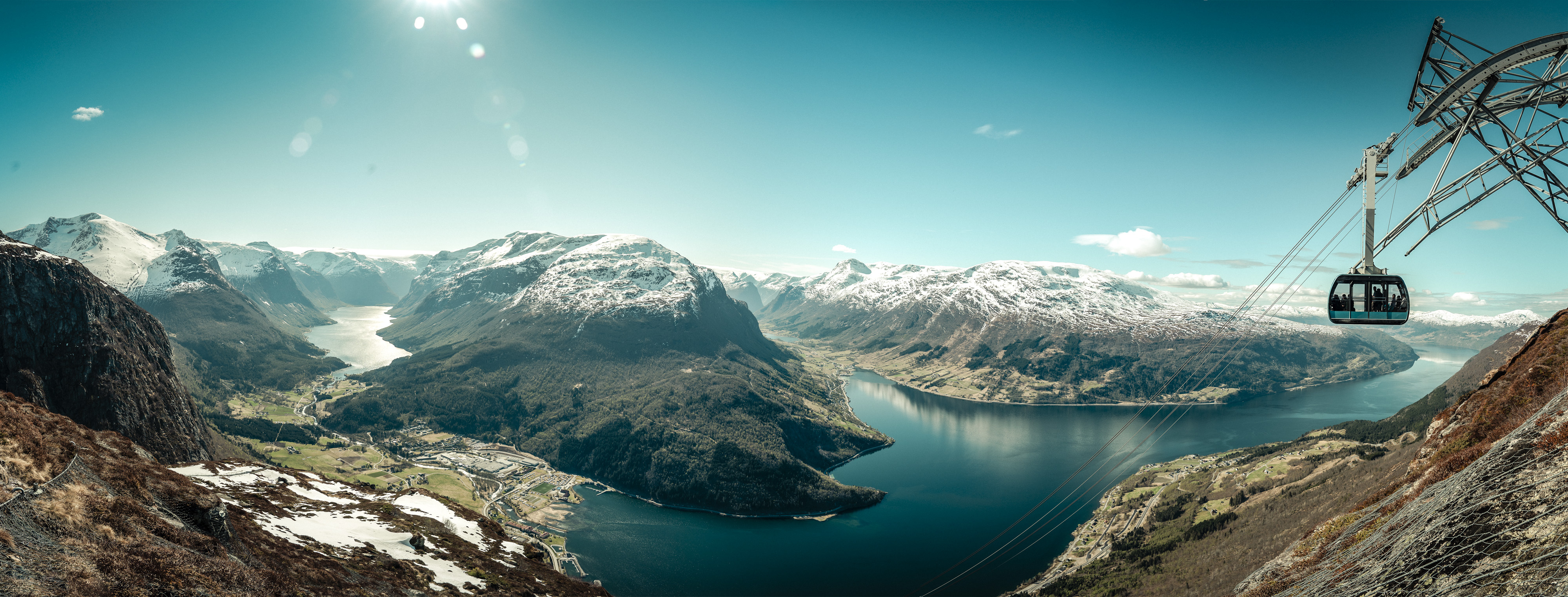 Et fjordlandskap med dype daler og snøkledde fjell i solskinn, fotografert ovenfra. Innerst i fjorden er det et tettsted. Foto. 