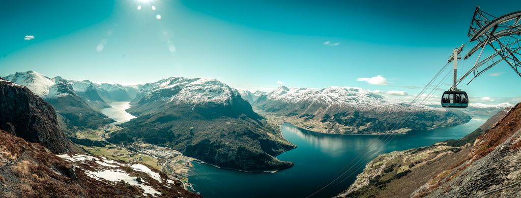 Gondol som kommer opp på fjellet til høyre. I bakgrunnen er et flott fjordlandskap med dype daler og snøkledde fjell. Foto. 