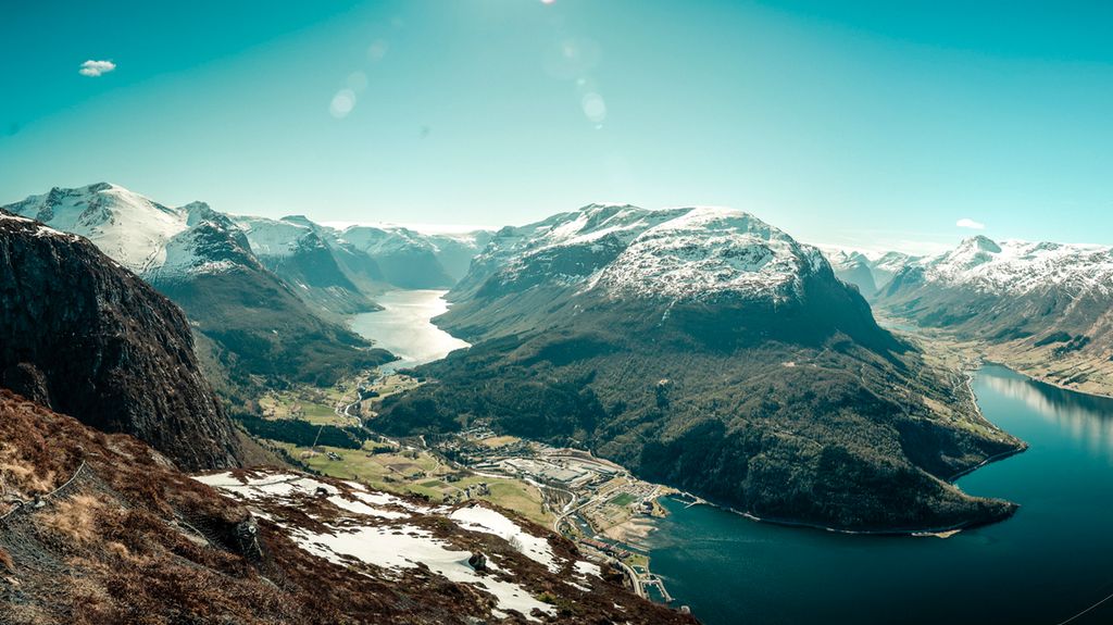 Et fjordlandskap med dype daler og snøkledde fjell i solskinn, fotografert ovenfra. Innerst i fjorden er det et tettsted. Foto.