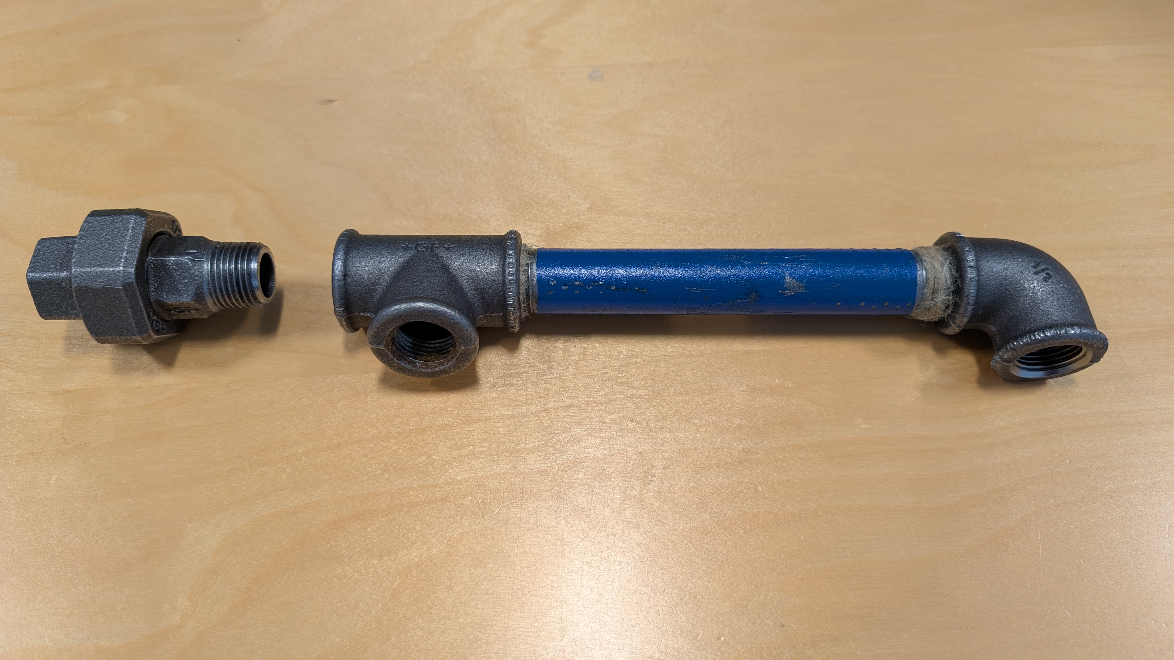 I rekkefølge fra venstre viser bildet en tippunion, et T-rør, et blått rør på cirka 15 centimeter og et albueledd. Både T-røret og albuen er skrudd på røret, som har gjenger i begge ender. Foto.