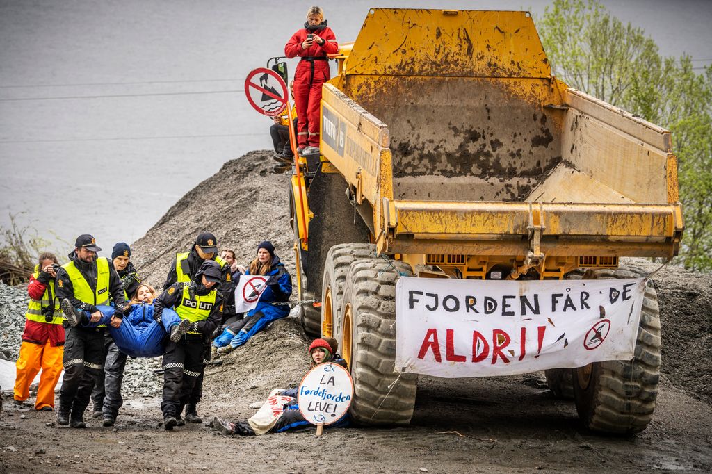 Til høyre står en stor dumper med et banner festet over bakhjulene. På banneret står det "Fjorden får de aldri!!". Demonstranter sitter ved dumperen. De har skilt der det står "La Førdefjorden leve!". Til venstre bærer fire politifolk en person bort fra området. Foto. 