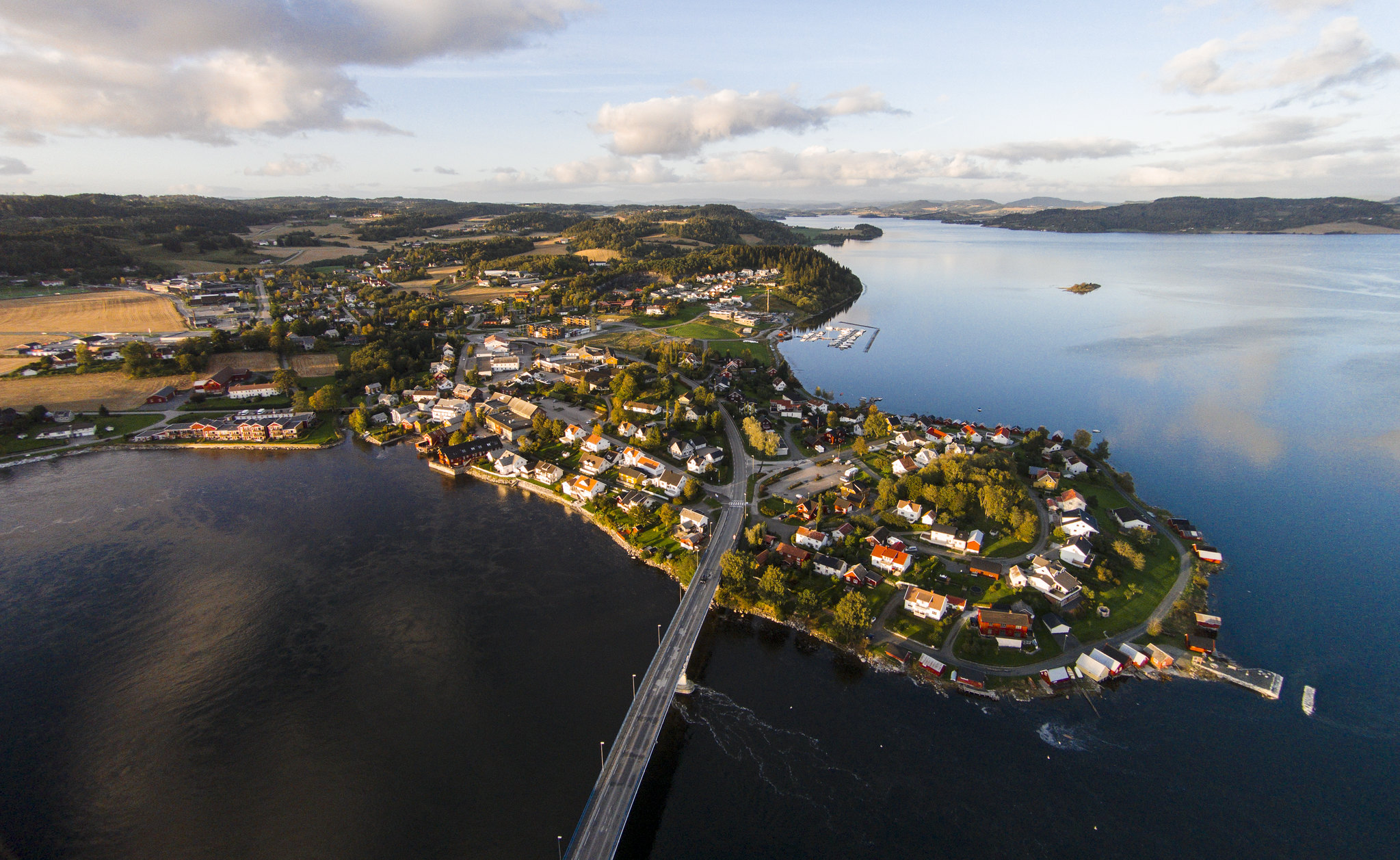 Et nes med bebyggelse stikker ut i en fjord. Ei bro går over til neset, og i bakgrunnen er det mer bebyggelse, skogkledde områder og noen åkre. Bildet er tatt fra lufta. Foto.