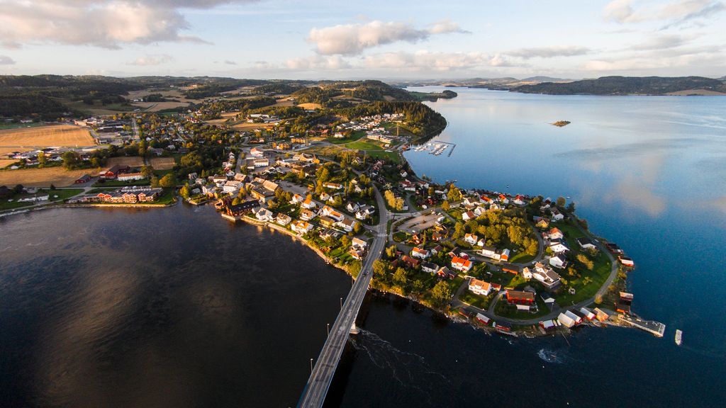 Et nes med bebyggelse stikker ut i en fjord. Ei bro går over til neset, og i bakgrunnen er det mer bebyggelse, skogkledde områder og noen åkre. Bildet er tatt fra lufta. Foto.