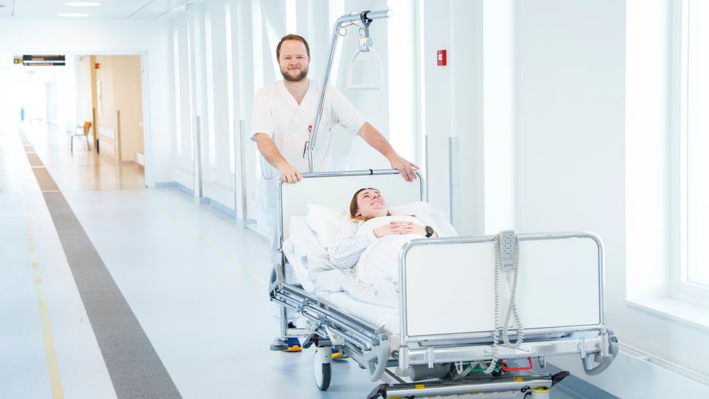 Person i uniform for helsepersonell står ved hodeenden av ei seng med en pasient i sykehuskorridor. Foto.