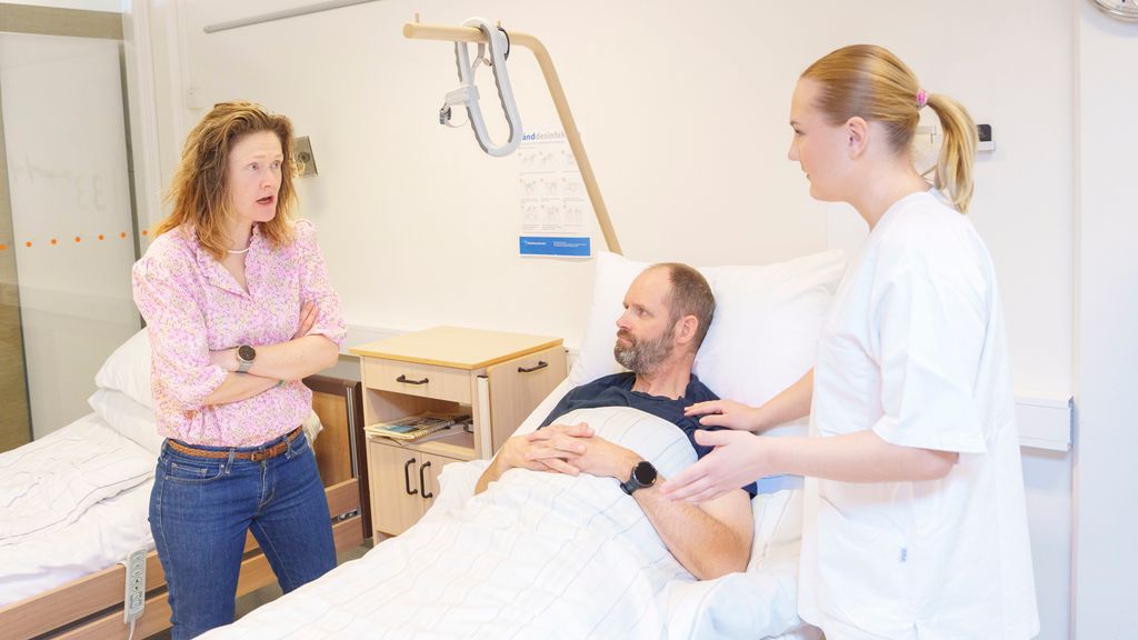 Helsefagarbeider i diskusjon med en pårørende ved en sengeliggende pasient. En kvinne i sivile klær står ved siden av ei seng hvor det ligger en mannlig pasient. Kvinnen har armene i kors, lener seg litt framover mot helsefagarbeideren som står på den andre siden av senga, og har et opprørt uttrykk i ansiktet mens hun snakker. Helsefagarbeideren holder den ene hånda på skulderen til mannen i senga og holder den andre armen litt ut til siden. Foto.