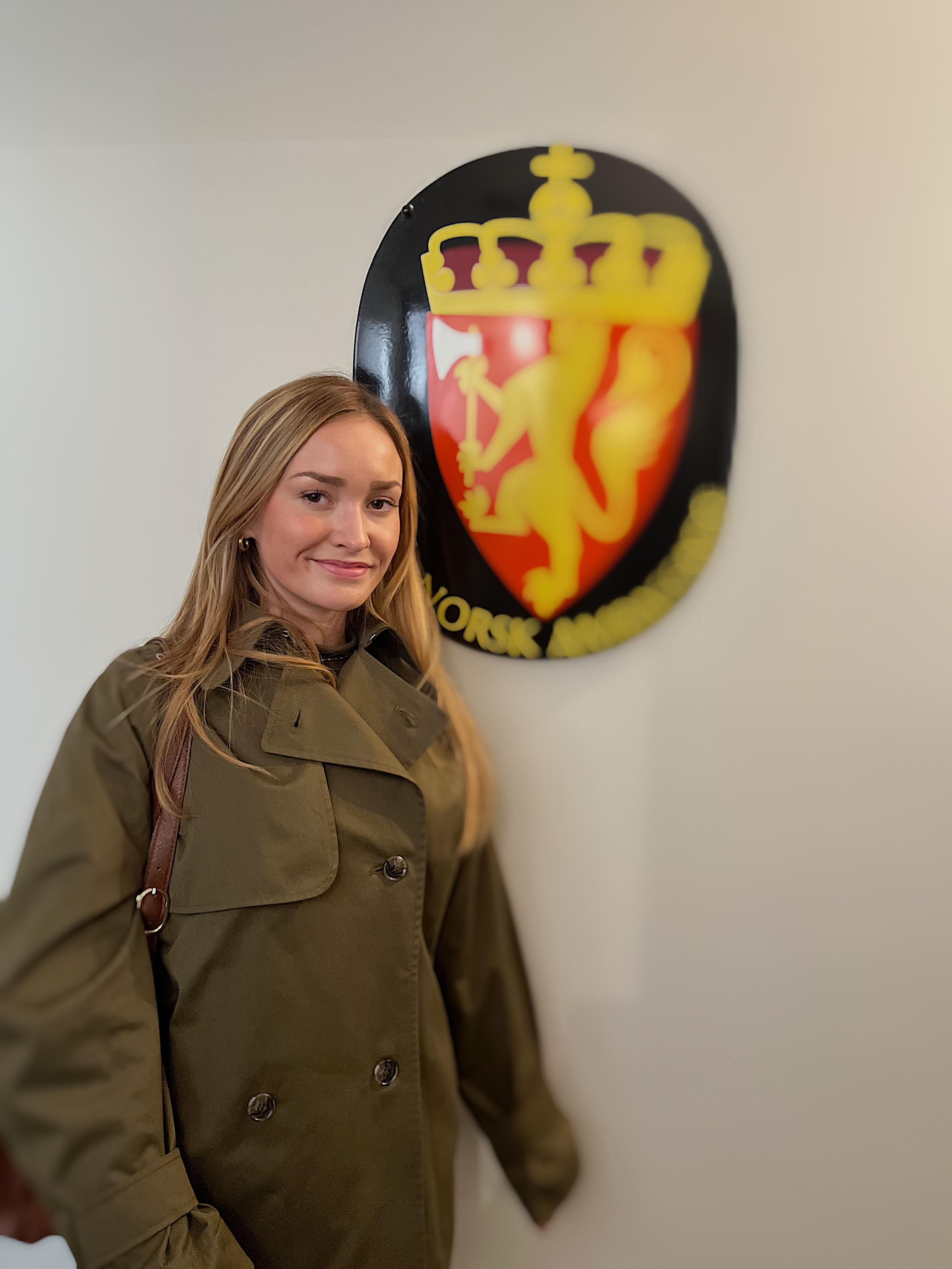 Smilande ung kvinne med mørkeblondt hår som har på seg ein grøn trenchcoat. I bakgrunnen det norske riksvåpenet. Foto.