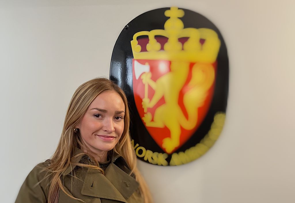 Smilande ung kvinne med mørkeblondt hår som har på seg ein grøn trenchcoat. I bakgrunnen det norske riksvåpenet. Foto.