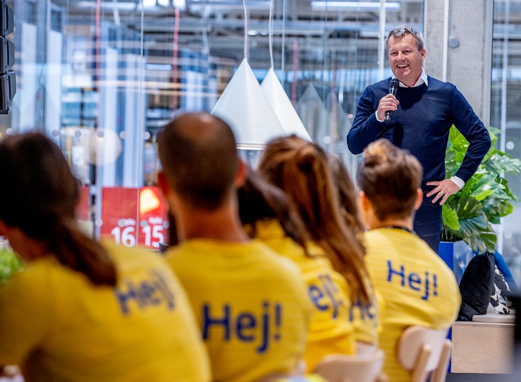 Person med mikrofon står smilende foran ei gruppe personer. De som hører på, sitter ved et bord og har på seg gule t-skjorter med påskriften "Hej". Foto.