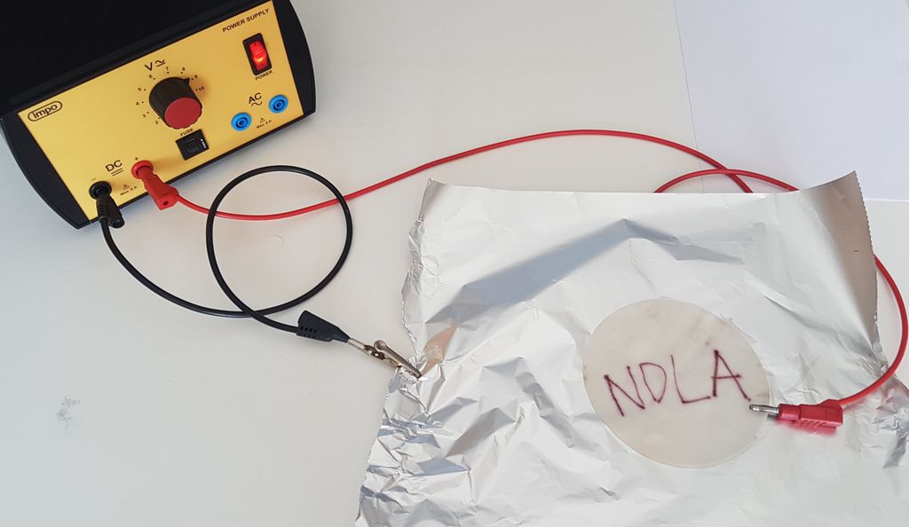 Aluminiumsfolie er koplet til en spenningskilde med en svart og en rød ledning. Fuktig papir med påskriften NDLA ligger oppå aluminiumsfolien. Foto.