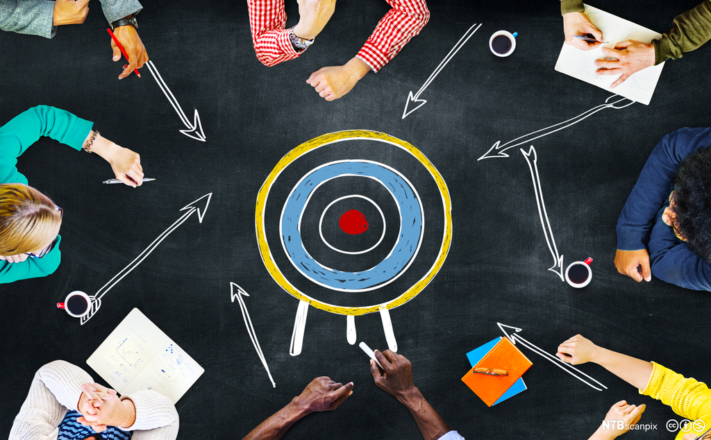 Fotografi fra fugleperspektiv ned på ei gruppe med personer som samarbeider. De former en ring, og i midten av den er det tegnet en rund blink med et rødt midtpunkt. Foto.