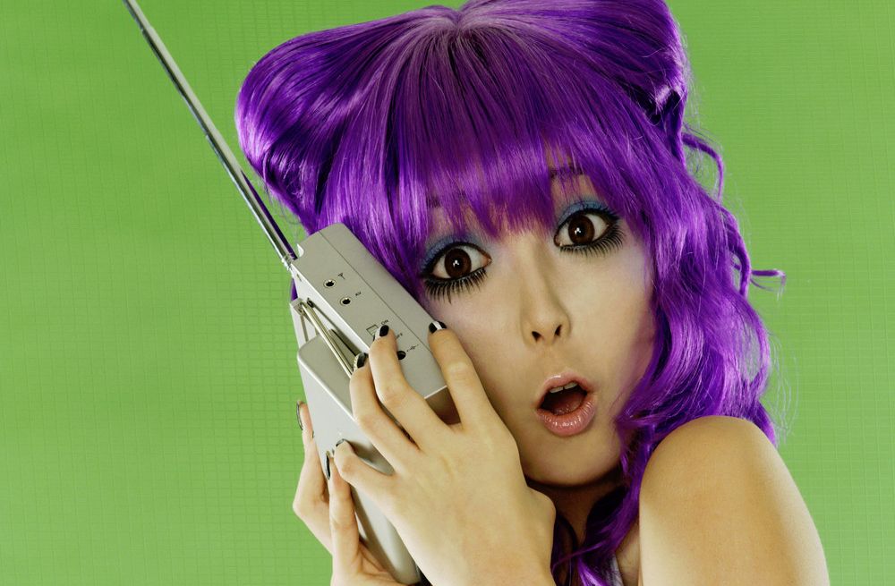 Ei jente med lilla hår holder en radio opp til øret og har et overrasket ansiktsuttrykk. Foto.