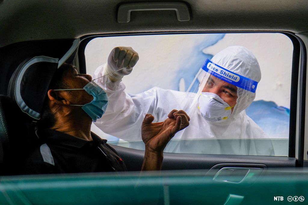 En maskert helsearbeider stikker en bomullspinne opp i nesen på en mann som sitter i en bil. Foto.