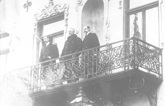 De tre kongene, Haakon 7. av Norge, Gustav 5. av Sverige og Christian 10. av Danmark, står i vinterfrakker og med hattene av oppe på en smijernsbalkong. Foto.