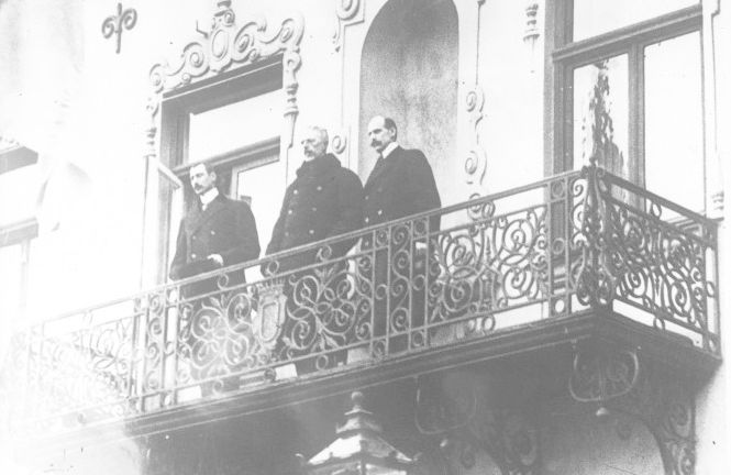Dei tre kongane, Haakon 7. av Noreg, Gustav 5. av Sverige og Christian 10. av Danmark, står i vinterfrakkar og med hattane av oppe på ein smijernsbalkong. Foto.
