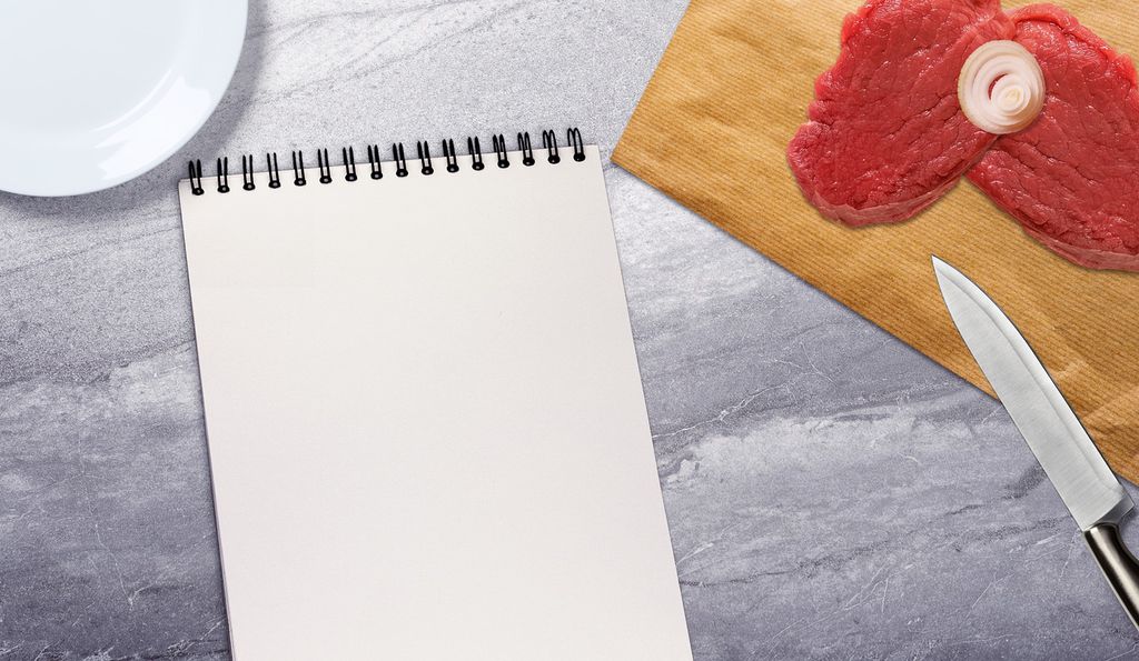To biffer på ei fjøl, kokkekniv og notatblokk på et bord. Foto.