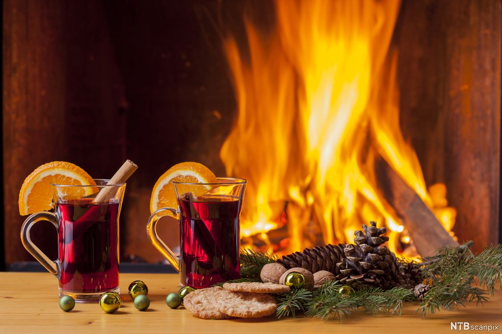 To glass med gløgg og småkaker i forgrunnen, peisbål i bakgrunnen. Foto.