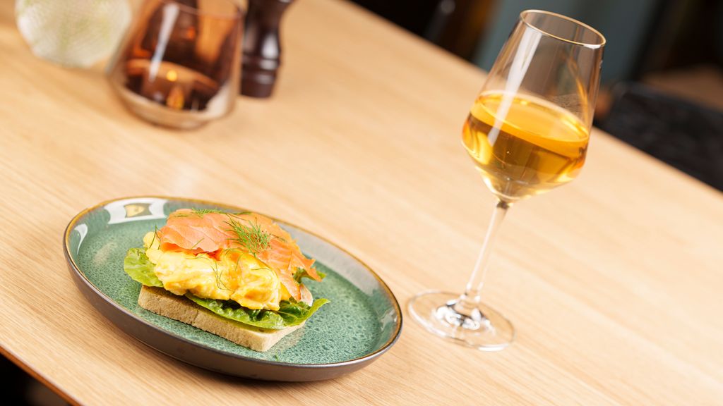Et smørbrød med eggerøre og røkelaks på en tallerken, Ved siden av står et vinglass fylt med sider. Foto.