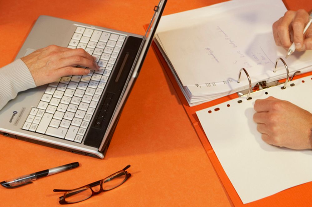 Nærbilde av hendene til noen som noterer på PC og på papir. Foto.