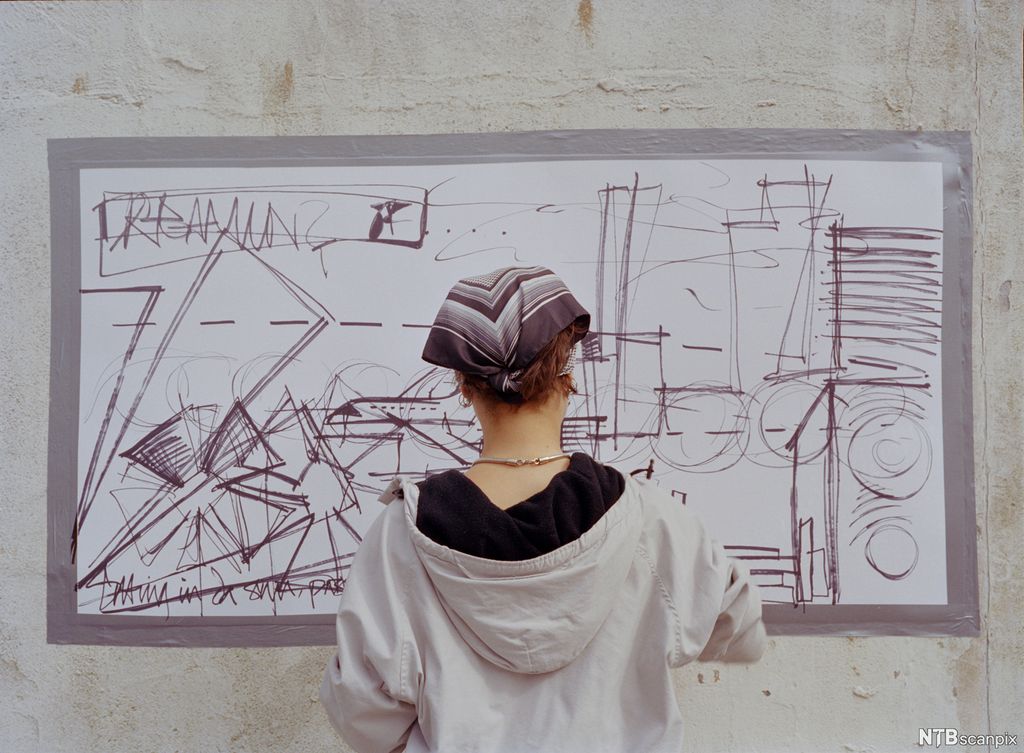 En jente med hettegenser og bandana tagger på en vegg. Foto. 