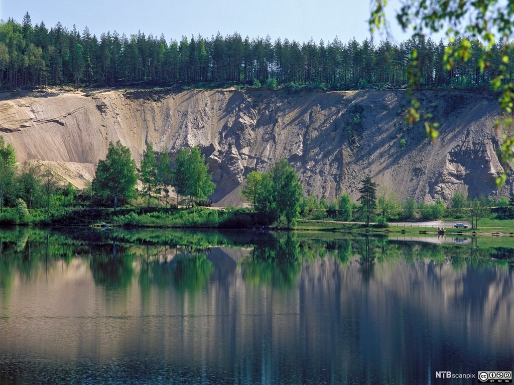 Stor skråning med sand og grus skråner ned mot et vann. Foto.