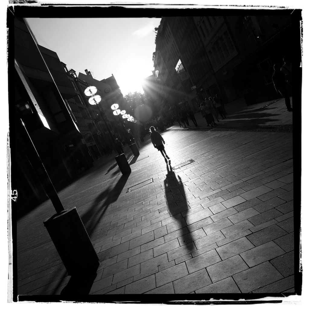 Svart-hvitt-bilde med sterke kontraster. En jente vandrer oppover en tom gate i silhuett mot sola. Foto.