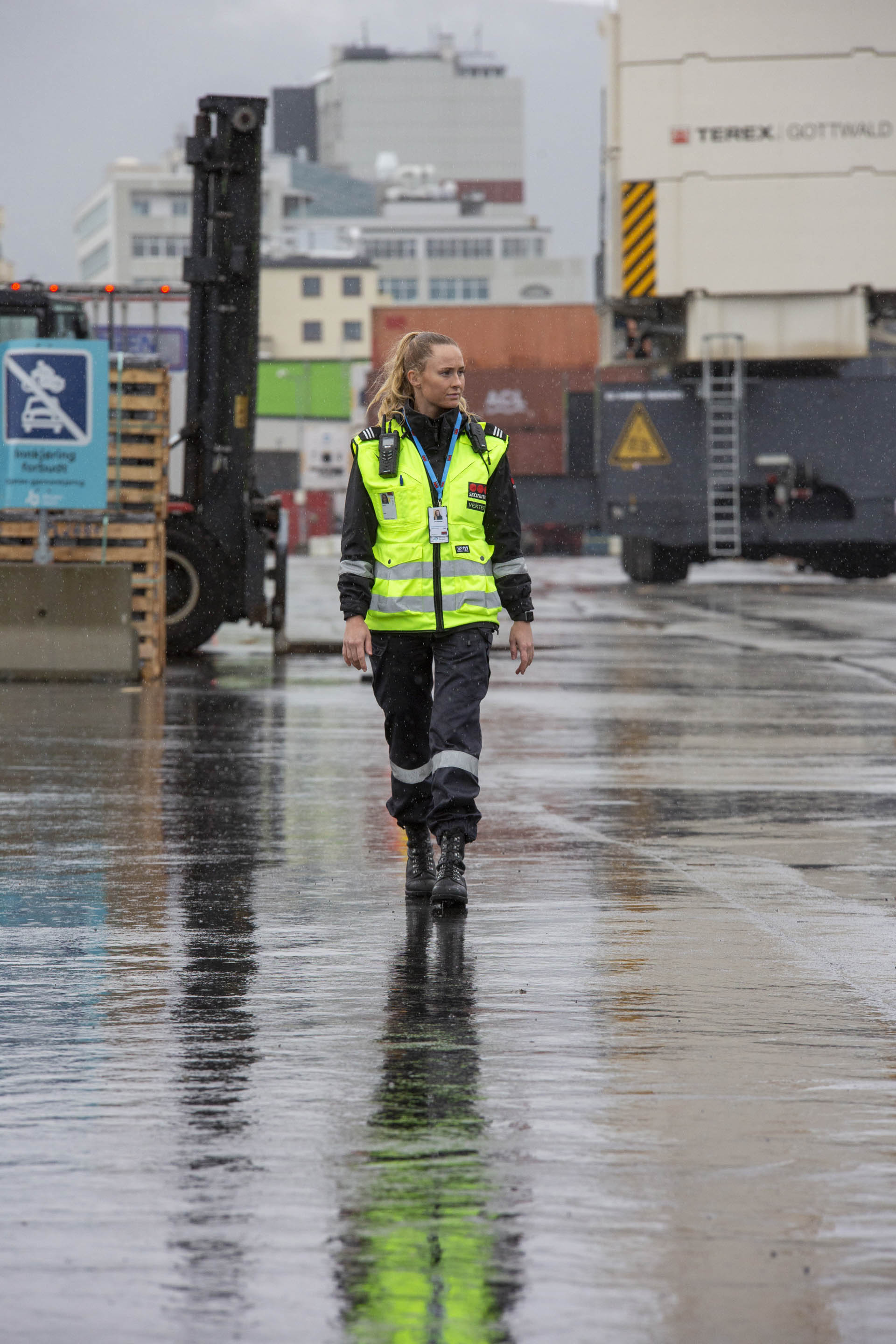 Kvinnelig vekter på inspeksjonsrunde i ei havn.  I bakgrunnen trailere,  en truck og lager- og administrasjonsbygg. Det regner. Foto.