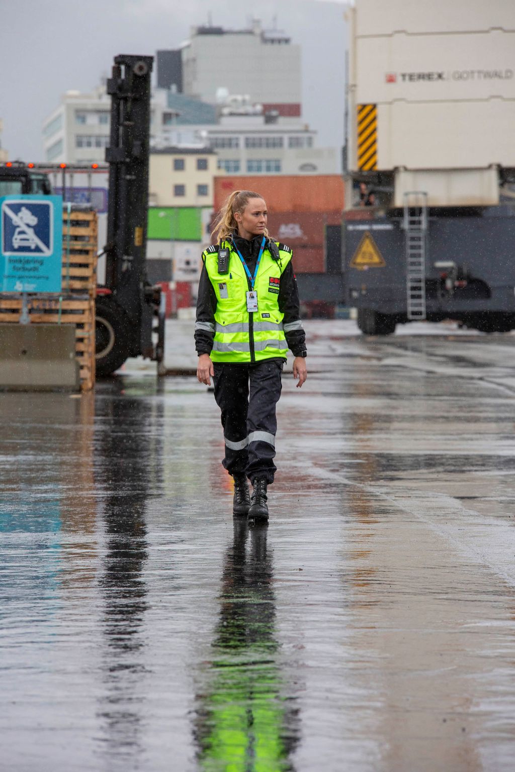 Ung kvinnelig vekter som går i et havneområde. Bak henne ses industribygg, konteinere og en truck. Foto.