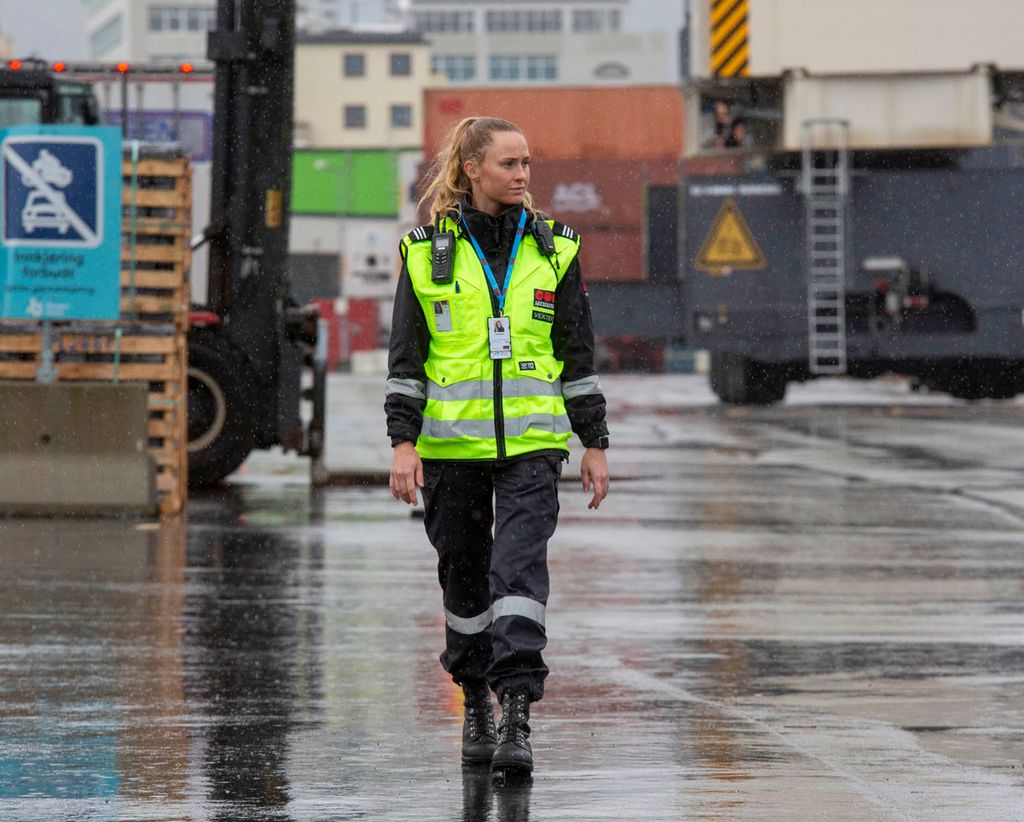 Kvinneleg vektar på inspeksjonsrunde i ei hamn. I bakgrunnen trailerar, ein truck og fleire lager- og administrasjonsbygg. Det regner. Foto.
