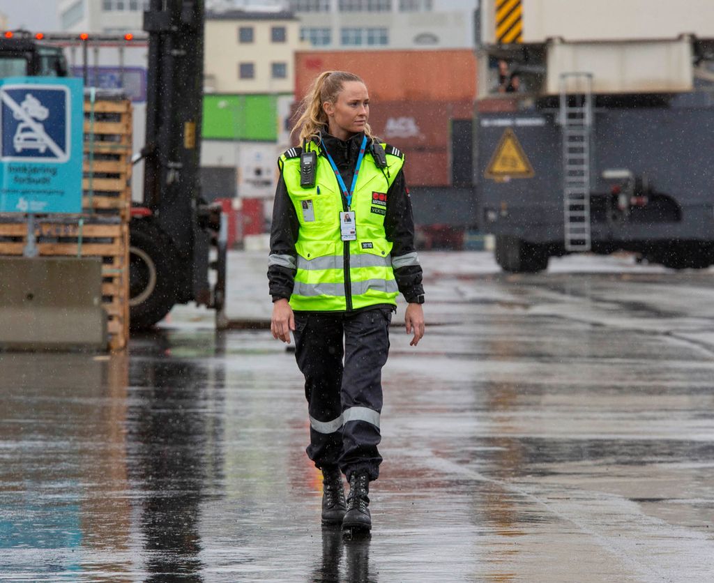 Kvinnelig vekter på inspeksjonsrunde i ei havn. I bakgrunnen trailere, en truck og lager- og administrasjonsbygg. Det regner. Foto.