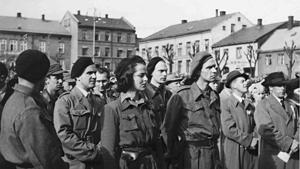 En rekke menn og ei kvinne står oppstilt i enkle militæruniformer. Hun er synlig bevæpnet. Foto. 