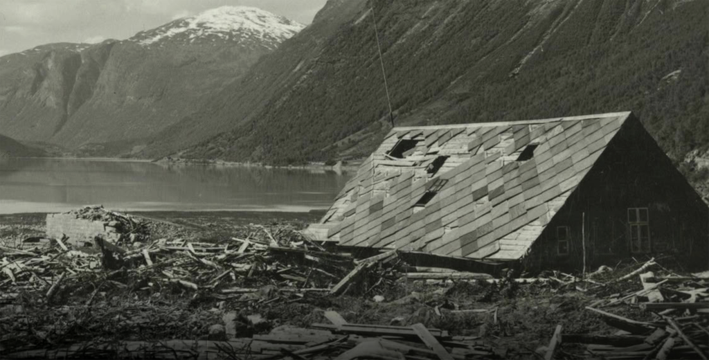 Et hus med ødelagt tak er omgitt av rasmasser og rester fra ødelagte bygninger. I bakgrunnen er restene av en mur, en innsjø og bratte fjell. Svart-hvitt-foto.