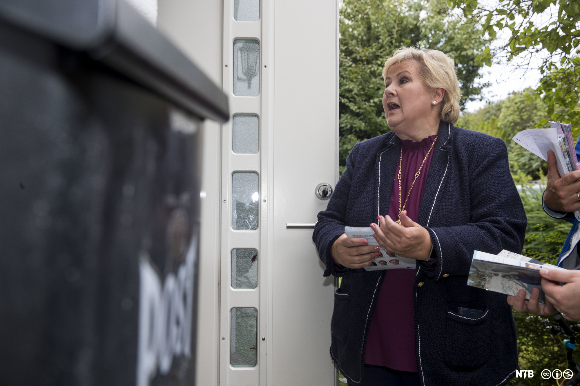 En kvinne med valgbrosjyrer i handa står ved inngangen til et hus og snakker med en person som står innafor døra. Personen er ikke synlig fordi det er plassert en stor mørk søppelkasse ved siden av døra. Foto.