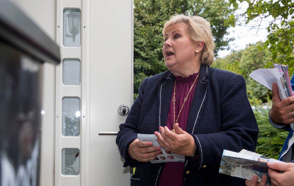 Ei kvinne med valbrosjyrar i handa står ved inngangen til eit hus og snakkar med ein person som står innanfor døra. Personen er ikkje synleg fordi det er plassert ei stor mørk søppelkasse ved sida av døra. Foto.