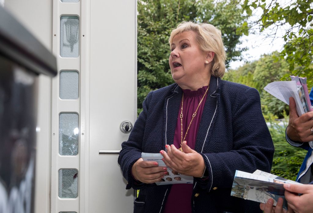 En kvinne med valgbrosjyrer i handa står ved inngangen til et hus og snakker med en person som står innafor døra. Personen er ikke synlig fordi det er plassert en stor mørk søppelkasse ved siden av døra. Foto.