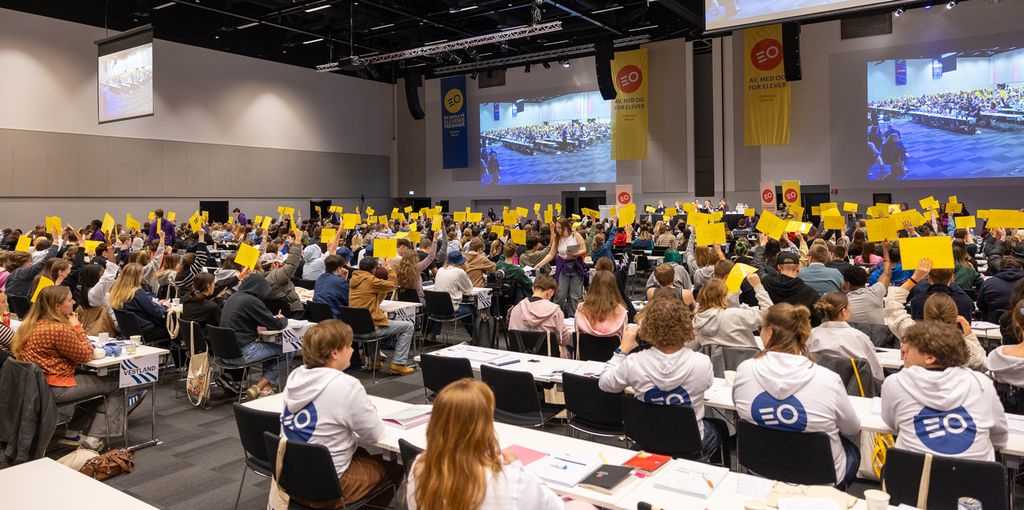 En stor forsamling ungdom sitter i en sal. Mange av dem holder opp et gult ark. Foto.