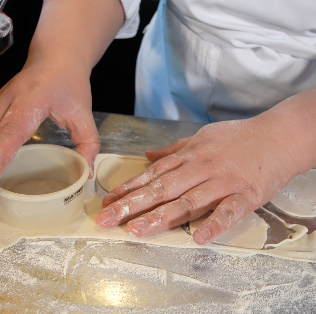 Nærbilde av tynn deig med mel rundt, en person bruker en utstikker til å stikke ut sirkler. Foto.