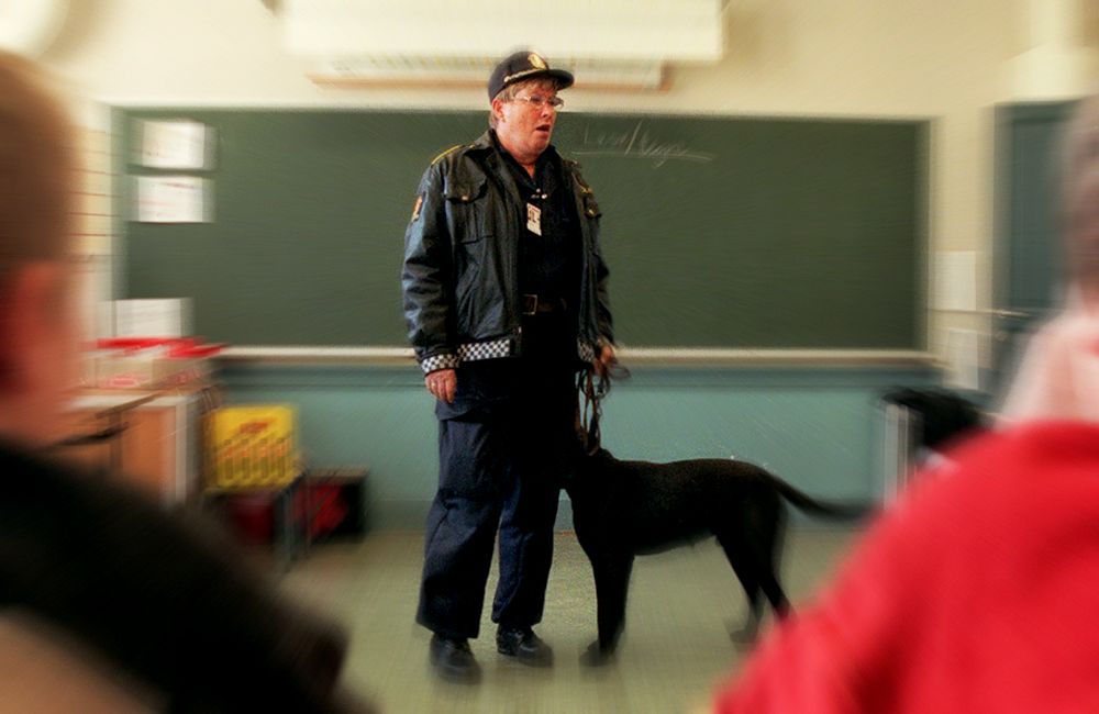 Politi i uniform står foran ei tavle i et klasserom og snakker. Foto.