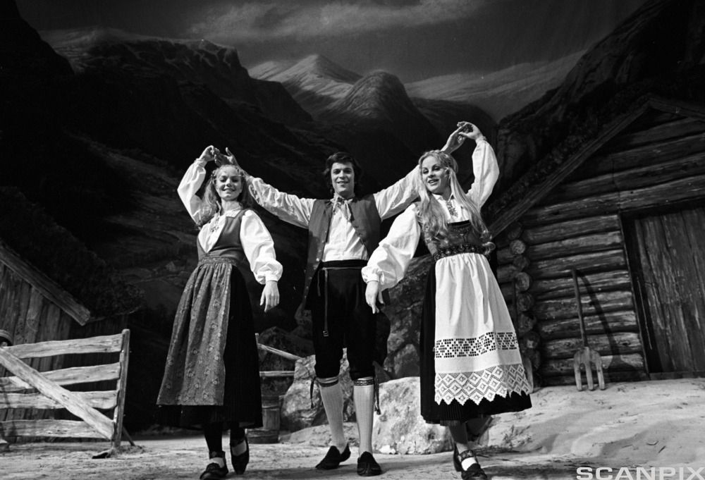 To kvinner og en mann holder hender foran en eldre fjellgård, ikledd norske bunader. Svart-hvitt-foto.