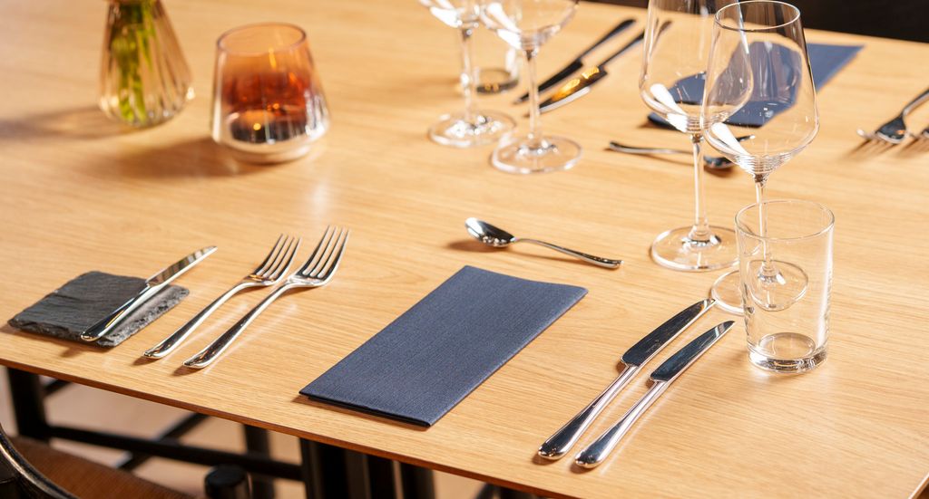 Et dekket bord i en restaurant. På bordet er det smørekniv, forrettsbestikk, hovedrettsbestikk, dessertskje, vinglass, vannglass og serviett. Foto.