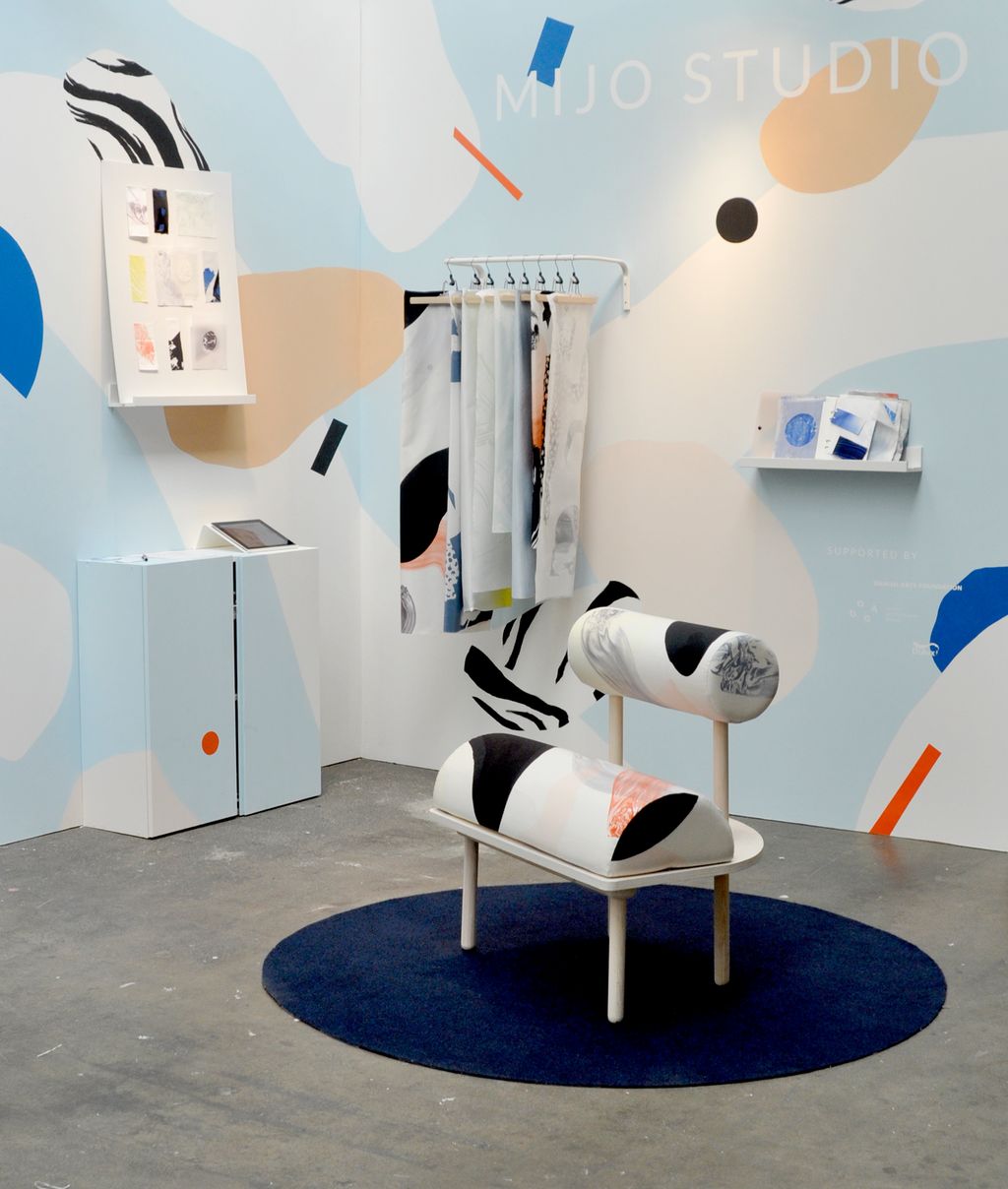 En messestand bestående av to vegger med fargerike former, noen tekstilprøver og et signaturmøbel oppå et rundt teppe. Foto.