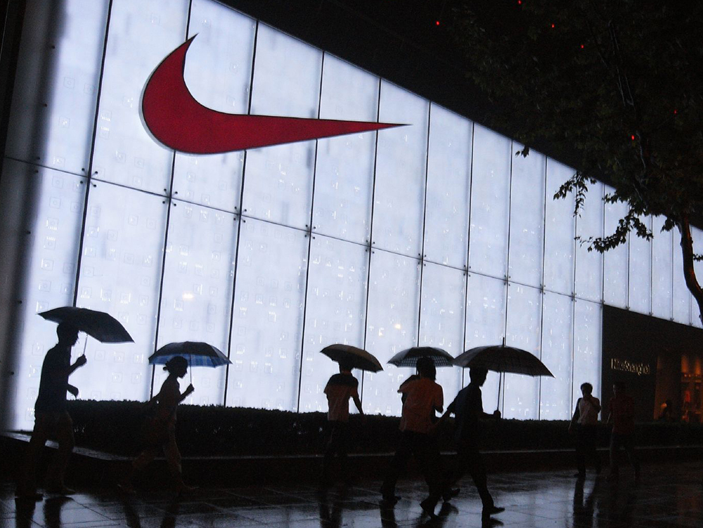 En stor Nike-logo på en opplyst yttervegg nattestid. Mennesker med paraply går forbi foran bygningen. Foto.