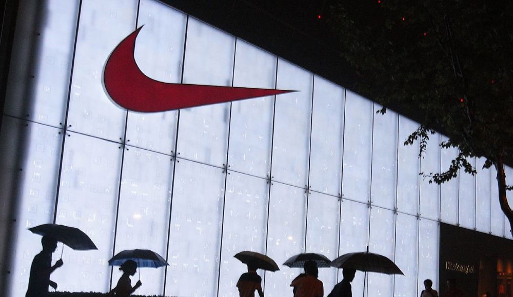 En stor Nike-logo på en opplyst yttervegg nattestid. Mennesker med paraply går forbi foran bygningen. Foto.